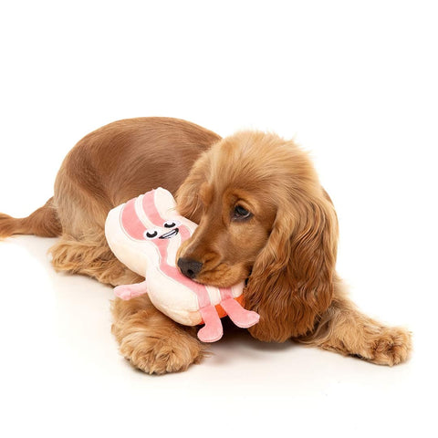Hond ligt met FuzzYard Meat Head Kevin Bacon – duurzaam pluche hondenspeeltje in de vorm van een plak bacon met lachend geborduurd gezicht en roze pootjes